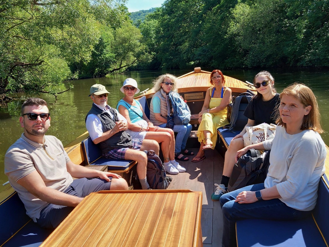 7 Personen sitzen in einem Boot. Im Hintergrund die Lahn und Bäume 