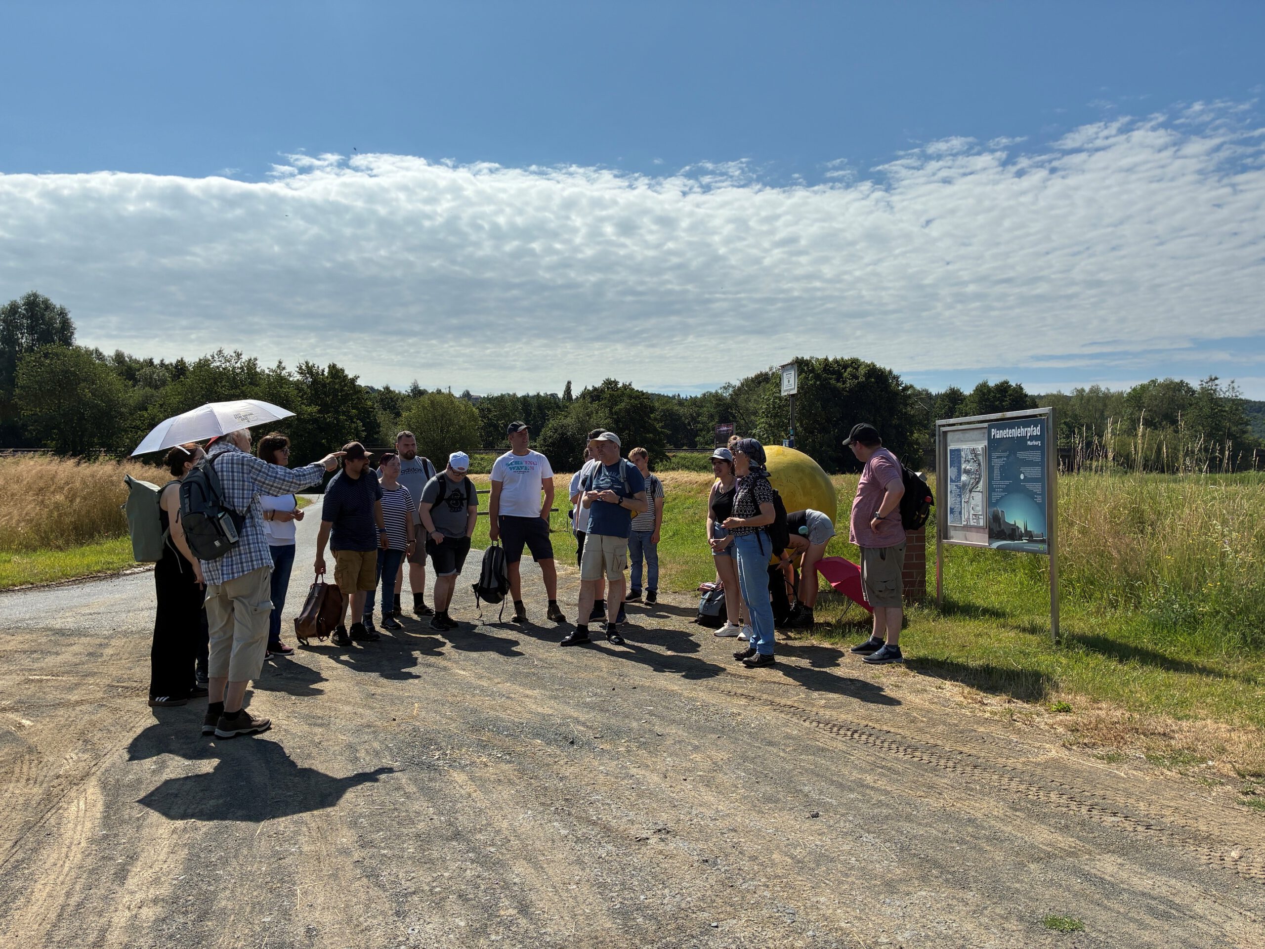 Wandergruppe mit Referent bei Station der Sonne, Wald und Wiese im Hintergrund 